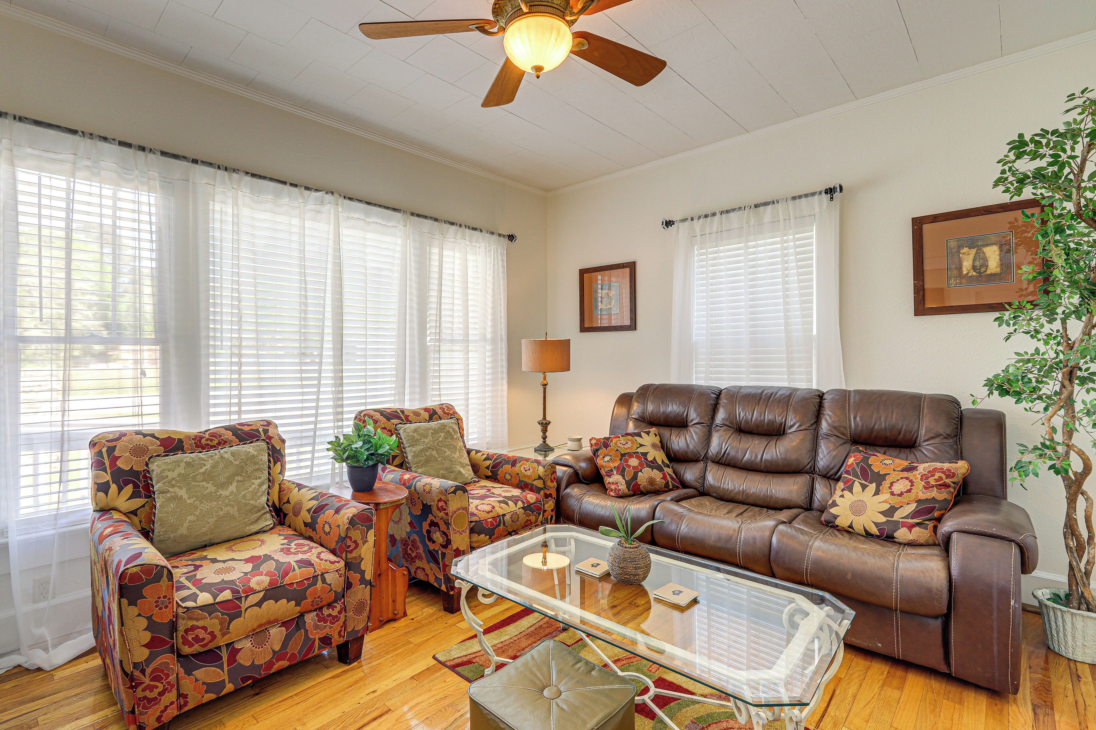 A living room with a brown leather sofa, two patterned armchairs, a glass coffee table, and a ceiling fan.