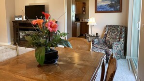A dining area with a wooden table, chairs, and a potted plant. A television is mounted on the wall, and a painting is visible. - Warm and Bright in the Heart of Downtown Friday Harbor (Friday Harbor)
