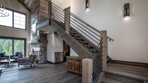 Amplia sala de estar con escalera de madera, chimenea de piedra y grandes ventanales con vistas al exterior.