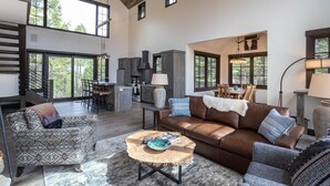 Un espacio habitable amplio con un sofá de cuero marrón, un sillón estampado y una mesa de centro redonda. Se ve en el fondo una zona de cocina con una cocina grande y una mesa de comedor.
