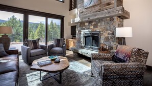 Un salón amplio con chimenea de piedra, asientos cómodos y ventanas grandes que ofrecen vistas al bosque.