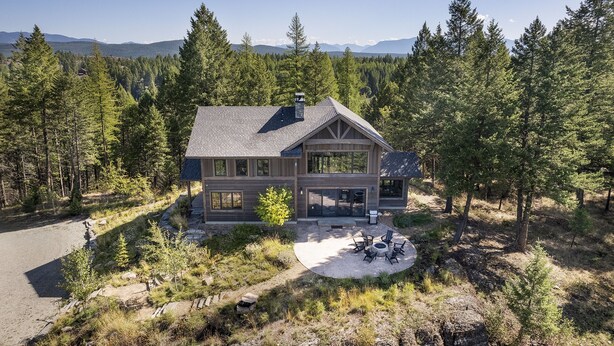Una casa moderna de dos pisos con una amplia terraza y un área con fogata, rodeada de un bosque denso y montañas al fondo.