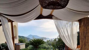 Mesa de cristal con fuentes de frutas y verduras, cortinas blancas y vistas a la vegetación y montañas.