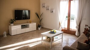 Una sala de estar con una televisión de pantalla plana, una mesa de centro blanca, un sofá y un ventanal con cortinas.