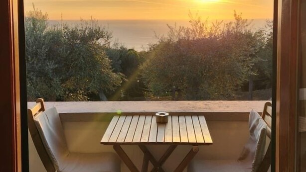 Un balcón con mesa de madera y dos sillas, con vistas a la puesta de sol sobre un paisaje costero.