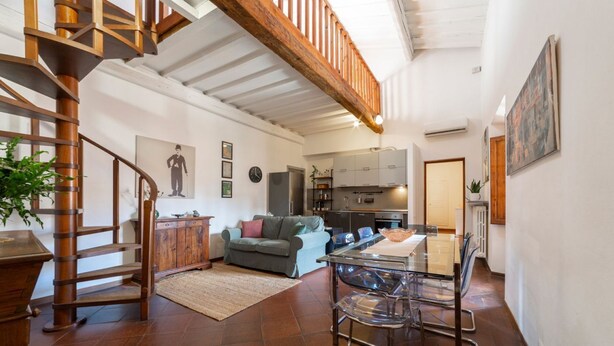A spacious living area with a wooden staircase, a dining table with chairs, a sofa, and a kitchen in the background.