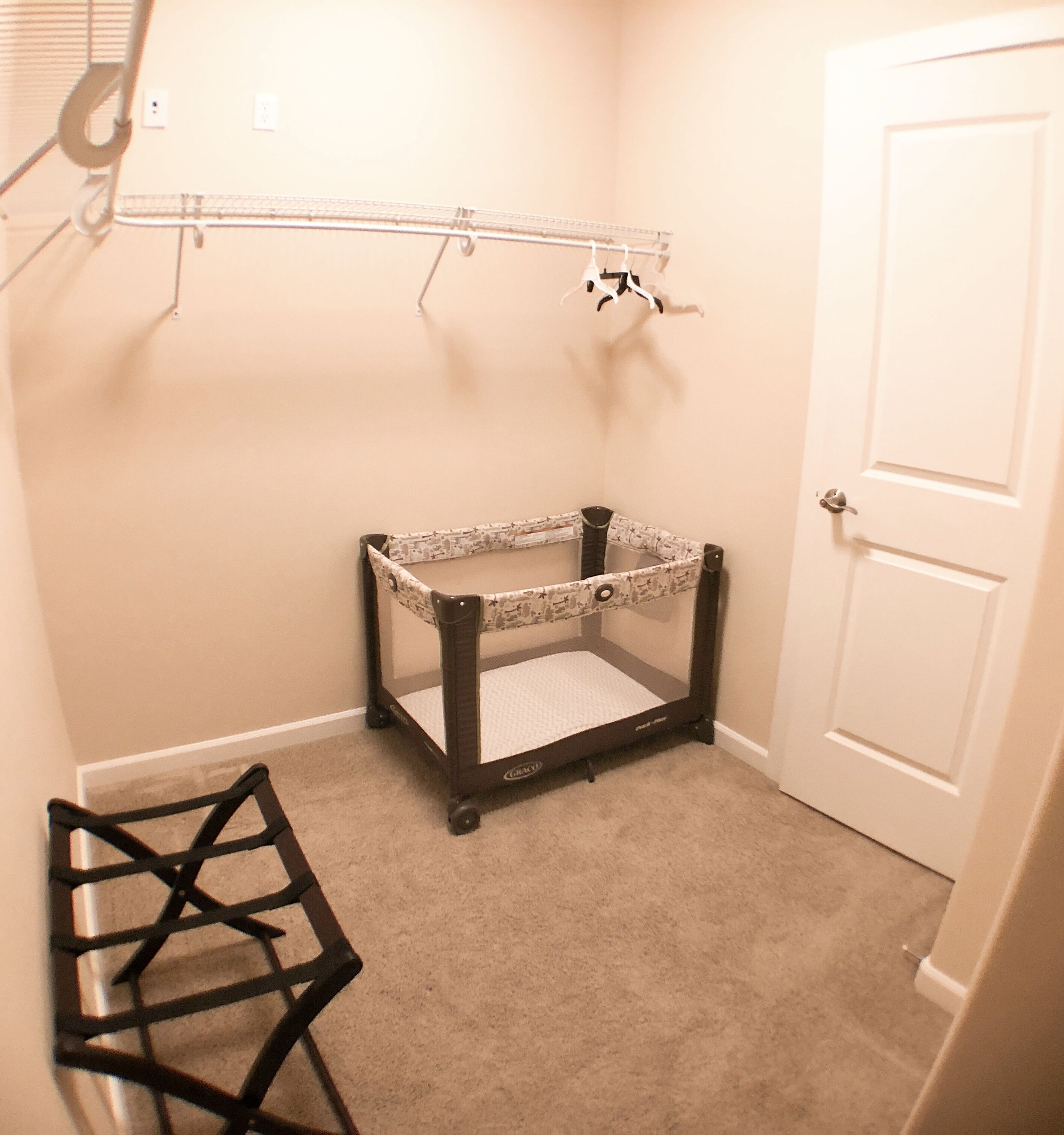 A baby crib with a patterned canopy, a small chair, and a coat rack in a room with beige walls and a carpeted floor.