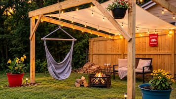 A wooden pergola with string lights, a hanging chair, a fire pit, and outdoor seating.