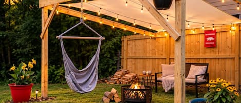 Una pérgola de madera con luces colgantes, un sillón hamaca, un hogar para fuego y asientos exteriores.