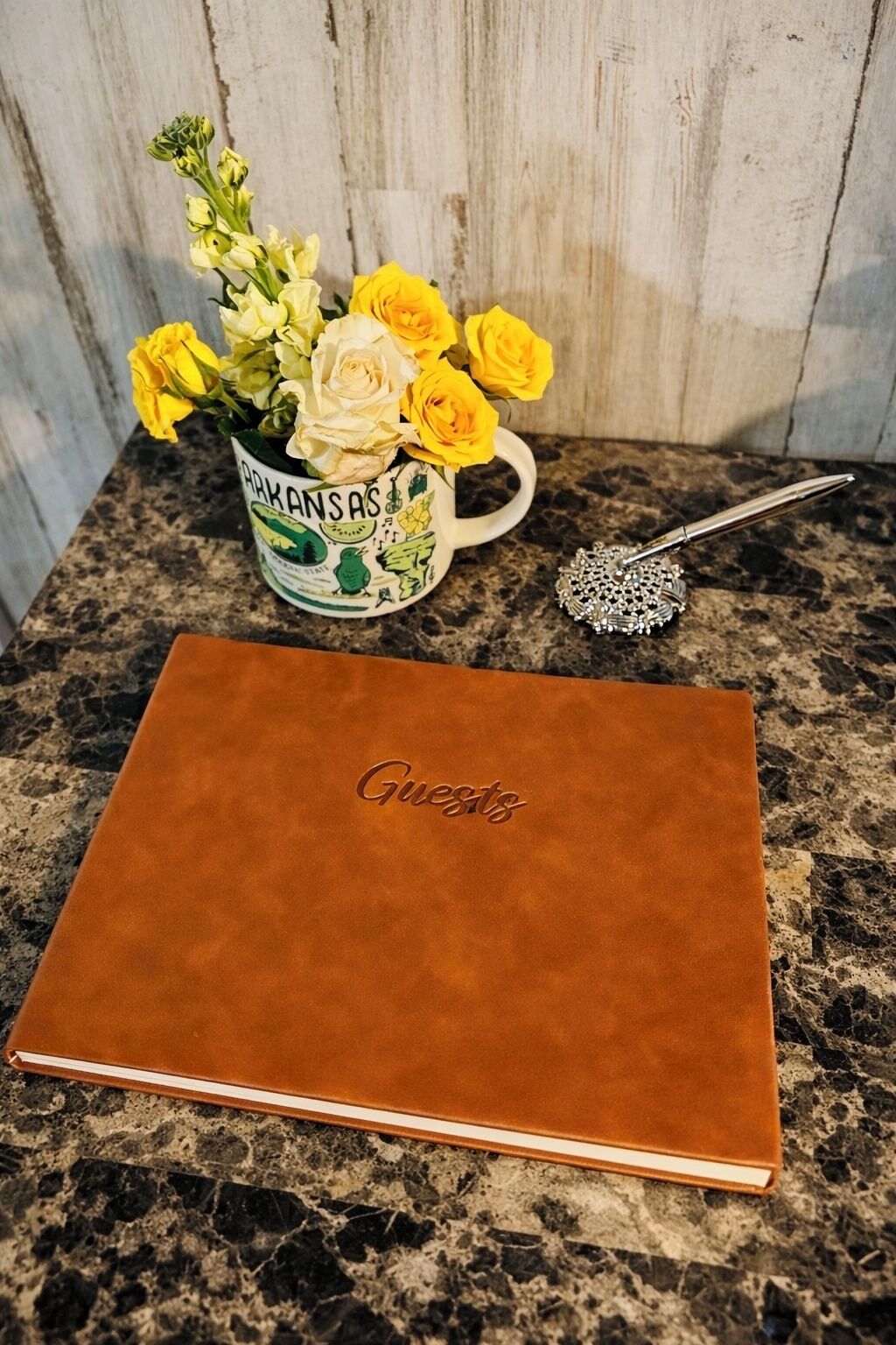 Un carnet en cuir marron avec le mot « Invités » en relief sur la couverture, une tasse ornée de motifs floraux et une cuillère décorative posée sur un comptoir en granit.