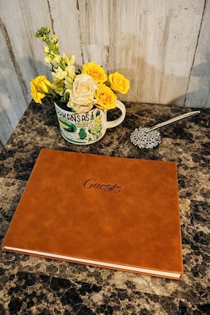 A brown leather notebook with "Guests" embossed on the cover, a mug with a floral design, and a decorative spoon on a granite countertop.
