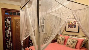 A four-poster canopy bed with a pink bedspread and palm tree pillows, a wooden door with a floral curtain, and a framed picture on the wall.