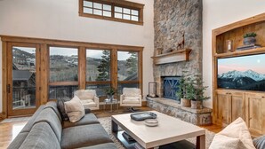 Un salon spacieux avec une cheminée en pierre, de grandes fenêtres offrant une vue sur un paysage montagneux et un coin salon confortable.