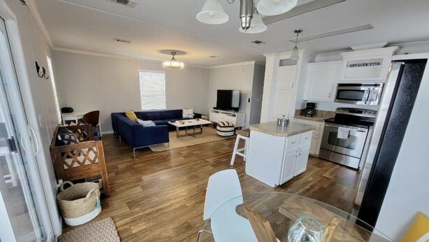 A modern living room with a sofa, a glass coffee table, and a TV. There is a kitchen area with a microwave, oven, and refrigerator.