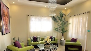 A spacious living room with a chandelier, green sofas, a glass coffee table, and a large potted plant.