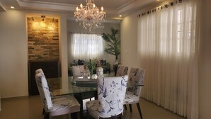 A dining area with a glass table, floral chairs, a chandelier, a stone wall, and a large window with curtains.
