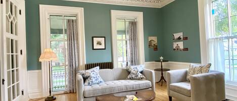 A living room with two sofas, a coffee table, and a side table. There are two windows with curtains, a radiator, and a lamp.