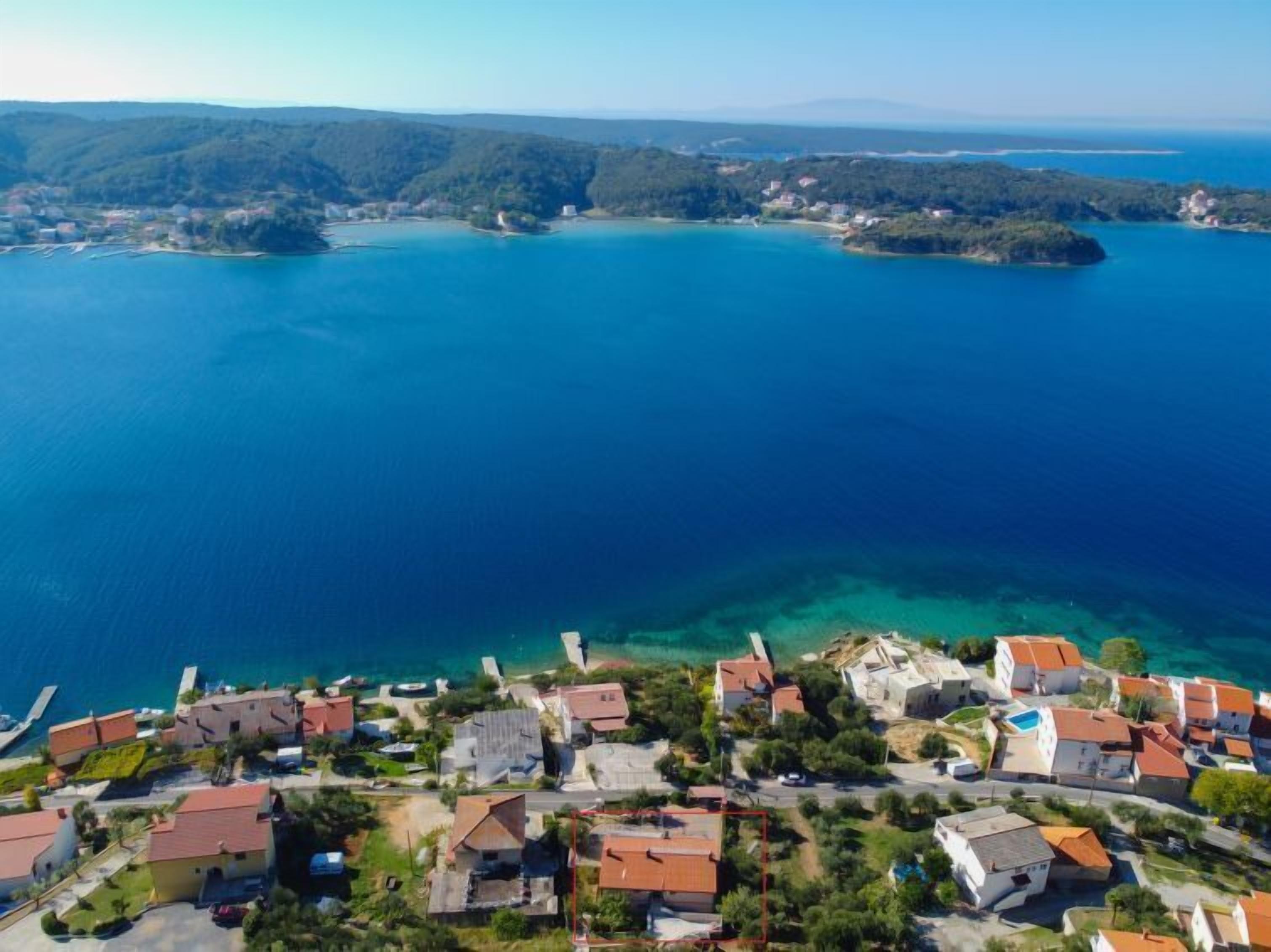 An aerial view of a coastal village with houses, clear blue sea, and hilly terrain in the background.