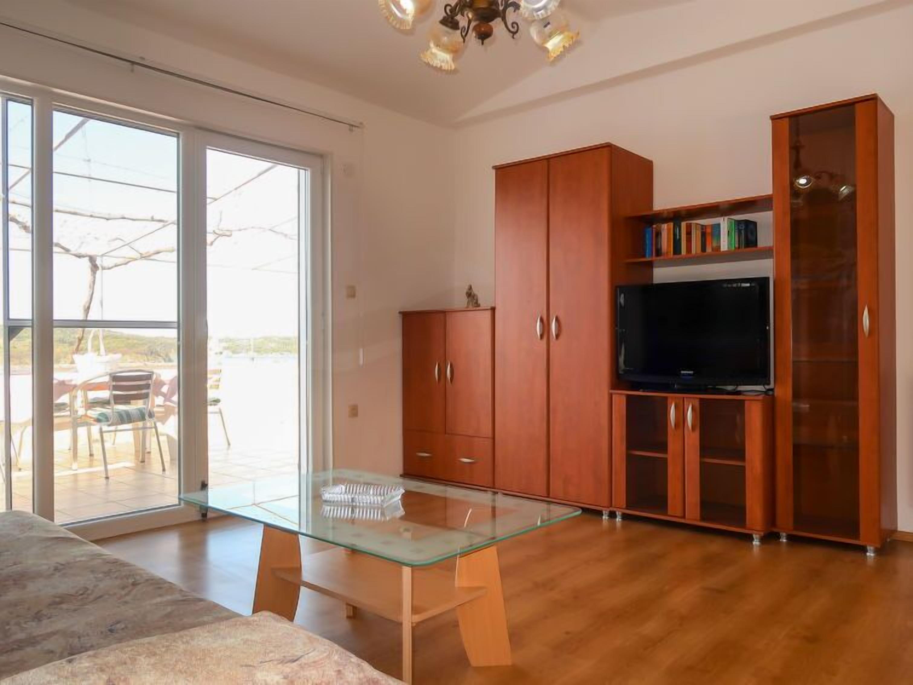 A modern living room with a glass-top coffee table, wooden cabinets, a flat-screen TV, and a sliding glass door leading to an outdoor area.