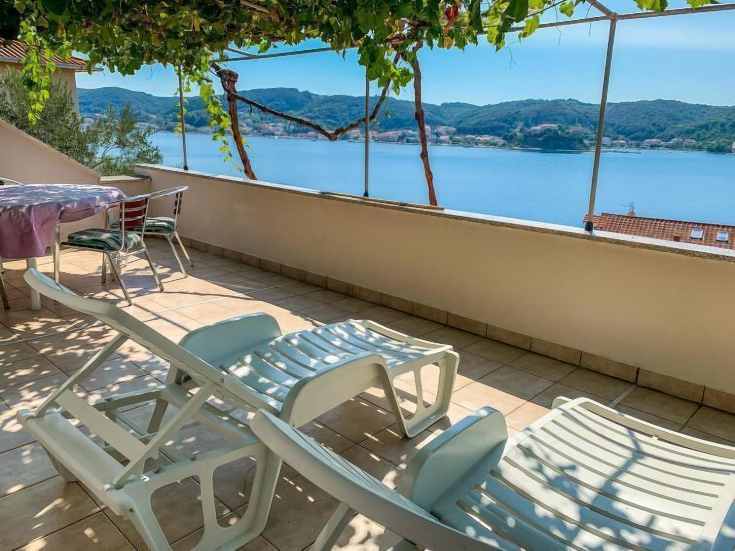 A terrace with white plastic chairs and a table, overlooking a lake with hills in the background.