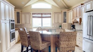 Cocina con una isla central, cuatro sillas de mimbre, hornos empotrados, refrigerador de acero inoxidable y un ventanal grande.