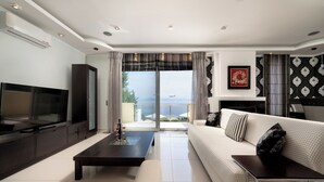 A modern living room with a large flat-screen TV, a dark coffee table, a white sofa with patterned cushions, and a view of the sea through a glass door. - Villa Alexandros luxury achillion corfu  (Corfu)