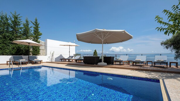 A swimming pool with lounge chairs and umbrellas, surrounded by a clear sky and trees. - Villa Alexandros luxury achillion corfu  (Corfu)