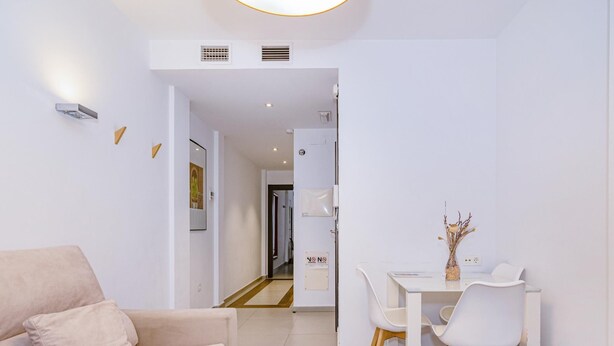 En moderne stue med en beige sofa, et spiseområde med et hvidt bord og stole, og en gang der fører til et andet værelse.