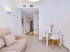 Ein modernes Wohnzimmer mit einem beige farbenen Sofa, einem Essbereich mit weißem Tisch und Stühlen und einem Flur, der in ein weiteres Zimmer führt.