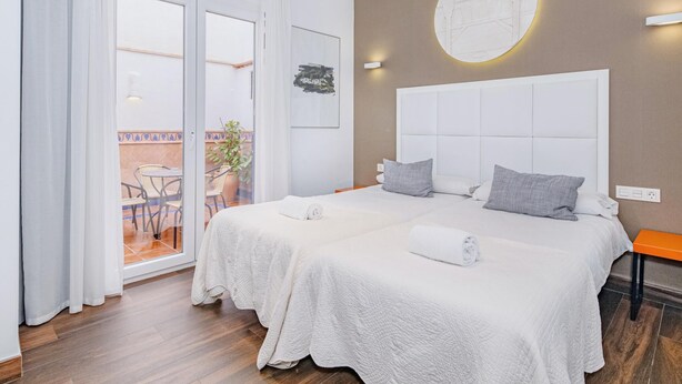 Un lit double avec une tête de lit blanche, une petite table de chevet orange et une vue sur un balcon agrémenté de chaises et d’une plante en pot.