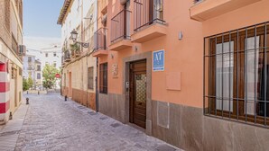Une ruelle étroite pavée, bordée d’immeubles, une grille métallique fermée et un panneau bleu sur lequel on peut lire « AT ».
