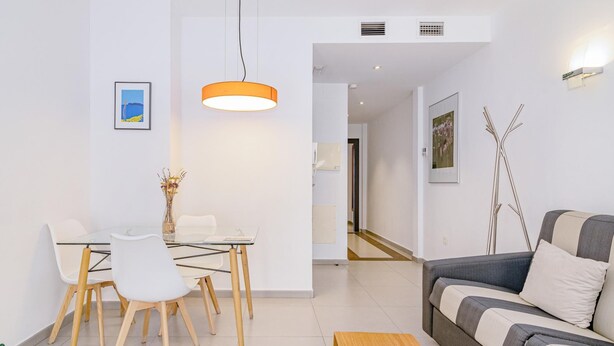 Una sala de estar moderna con un sofá, una mesa de comedor con sillas, una lámpara colgante y un pasillo que lleva a otra habitación.
