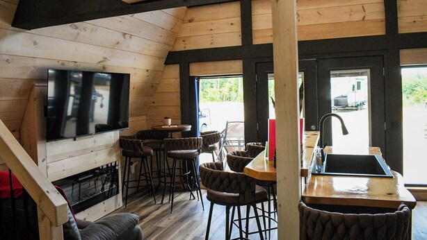 A cozy cabin interior with a wooden ceiling, a TV mounted on the wall, a bar area with stools, and a sink. - Picturesque Mountain Views, Direct Snowmobile & ATV trail access. 30 minutes to Sunday River and Black Mountain of Maine. (Andover)