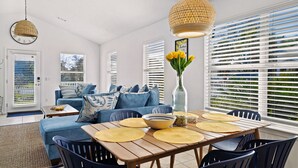 Een moderne woonkamer met een houten eettafel, een blauwe bank en een groot raam met zonwering.