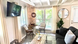 Ein Wohnzimmer mit einem Holztisch, einer schwarzen Couch und zwei Korbstühlen. Ein Fernseher ist an der Wand montiert, und es gibt ein Fenster mit Vorhängen.