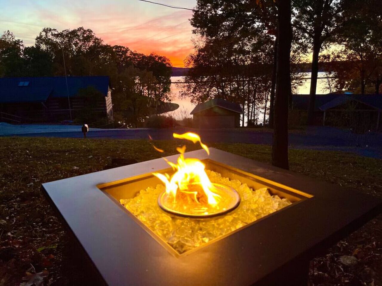 A square fire pit with a circular fire in the center, surrounded by a metal frame, set on a grassy area with trees and a sunset in the background.