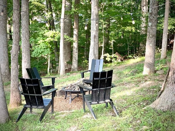 Dos sillas Adirondack alrededor de un pequeño hogar para fuego en un área boscosa.