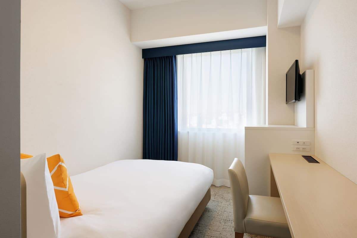 A hotel room with a bed, a desk, a chair, a television mounted on the wall, and a window with curtains.