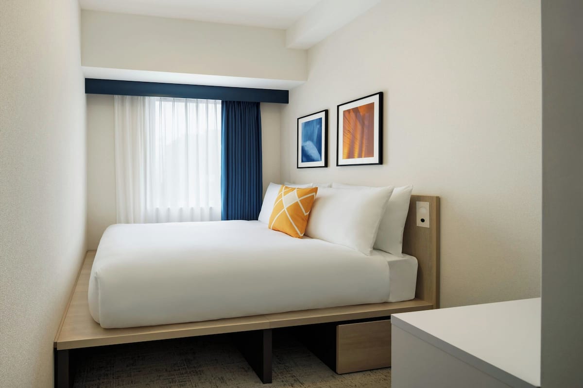 A hotel room with a bed, a bedside table, a window with curtains, and two framed pictures on the wall.