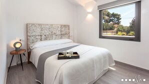 Un dormitorio moderno con una cama grande, una mesita de noche con lámpara y una ventana con vistas a árboles y edificios.