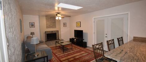 A living room with a sofa, TV, and fireplace, a dining area with chairs, and a kitchen with granite countertops.
