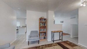 A spacious living area with a grey armchair, a wooden bookshelf filled with books, and a small wooden table. - NEW Renovated Private 3/2Home(King)Near Balloon Fiesta Park, Quiet Neighborhood! (Albuquerque)