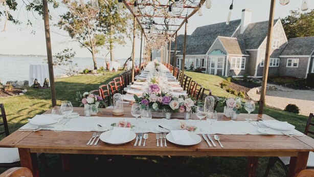 Una larga mesa de madera dispuesta para una comida, con manteles blancos, centros de mesa florales y sillas.