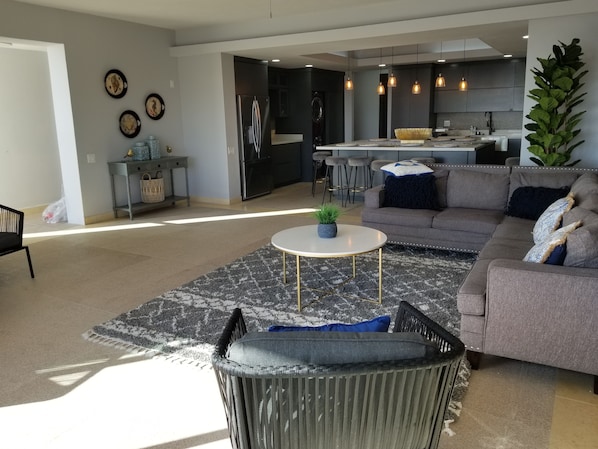 Un salon moderne avec un canapé gris, une table basse ronde et un fauteuil noir. On aperçoit, à l’arrière-plan, une cuisine avec des meubles sombres et un éclairage suspendu.