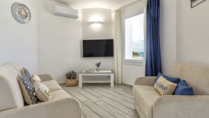 Ein Wohnzimmer mit zwei beigen Sofas, einem weißen Couchtisch, einem an der Wand montierten Flachbildfernseher und einem Fenster mit Vorhängen.