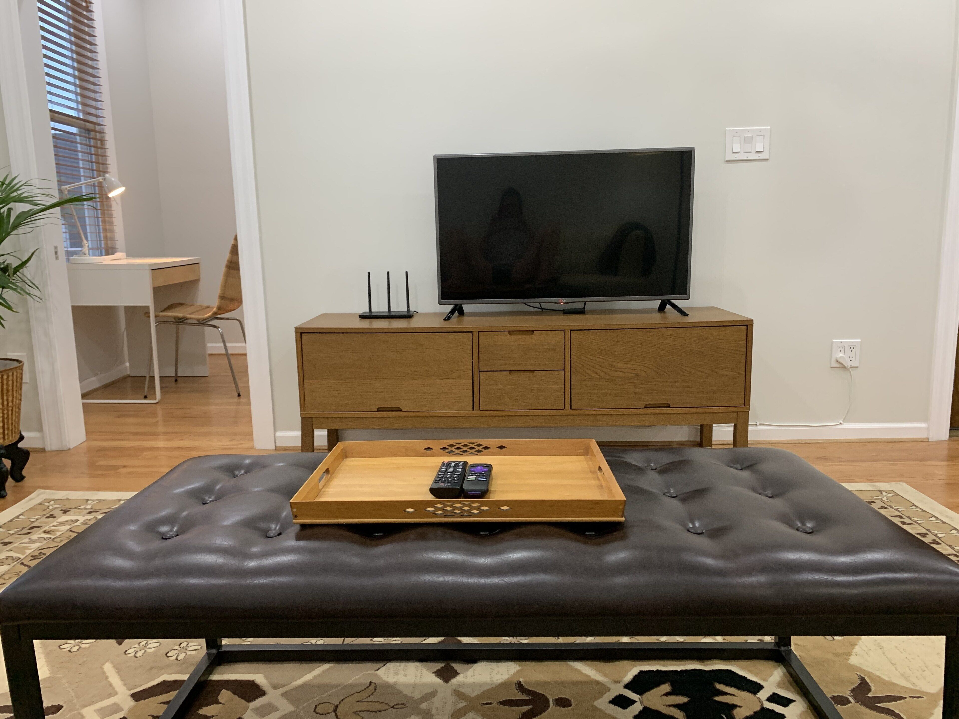 Una sala de estar con un reposapiés de cuero oscuro, una mesa de centro de madera con un control remoto, un televisor sobre un mueble de madera y un escritorio blanco con una silla.