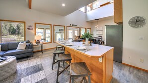 A modern kitchen with a central island, wooden bar stools, and a dining area with a sofa and coffee table. - Expansive Waterfront Escape w/ Kayaks + SUPs! (Sagle)