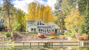 Une maison à deux étages avec une jetée s’avançant dans un lac paisible, entourée d’un feuillage automnal et d’un jardin bien entretenu.
