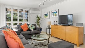 A modern living room with a grey sofa, orange cushions, a glass coffee table, a wooden cabinet, a flat-screen TV, and artwork on the wall. - Byron Breeze 7 ~ Luxury Penthouse 3 Bedroom Apartment (Byron Bay)