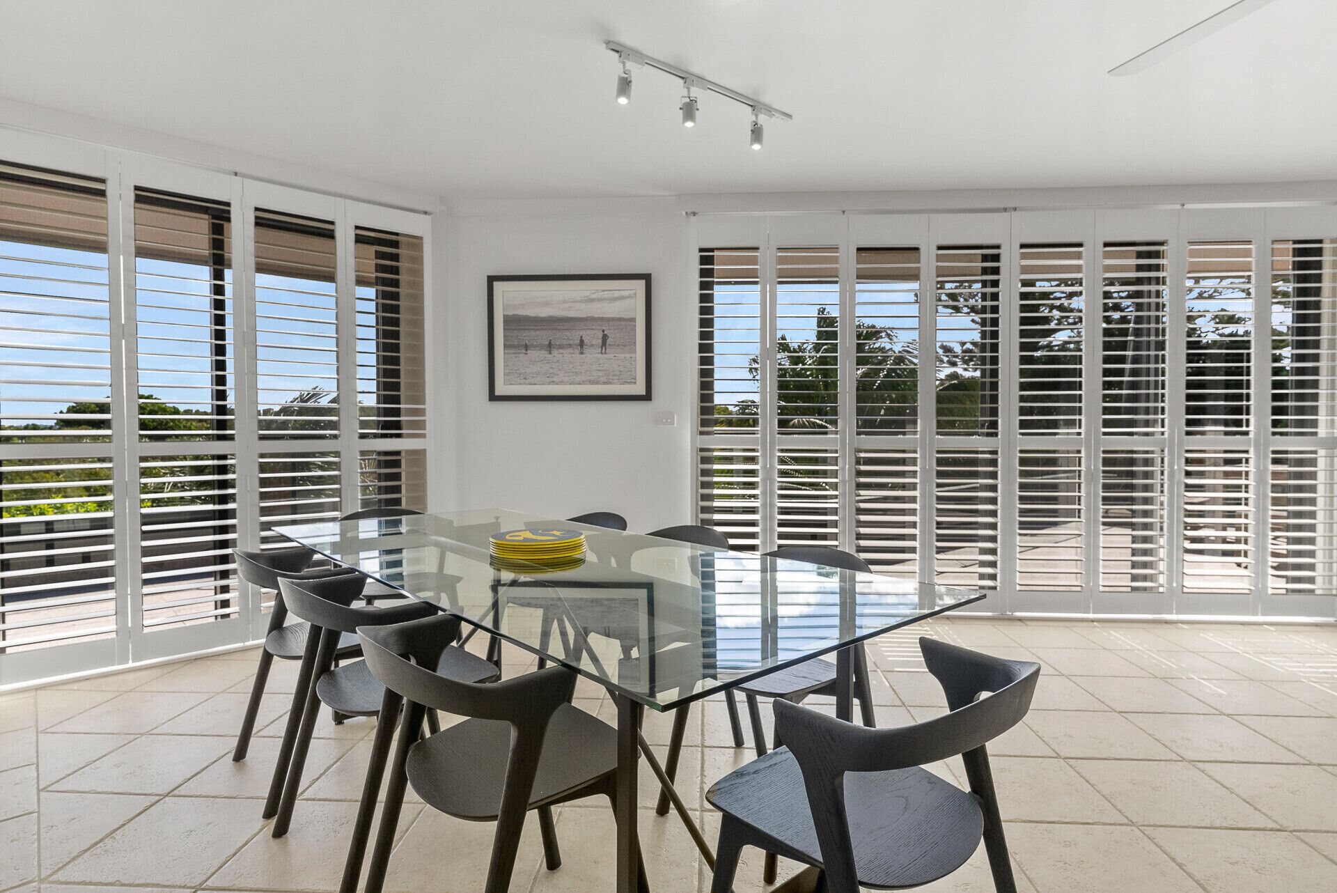 A modern dining area with a glass table and chairs, large windows with blinds, and a framed picture on the wall.